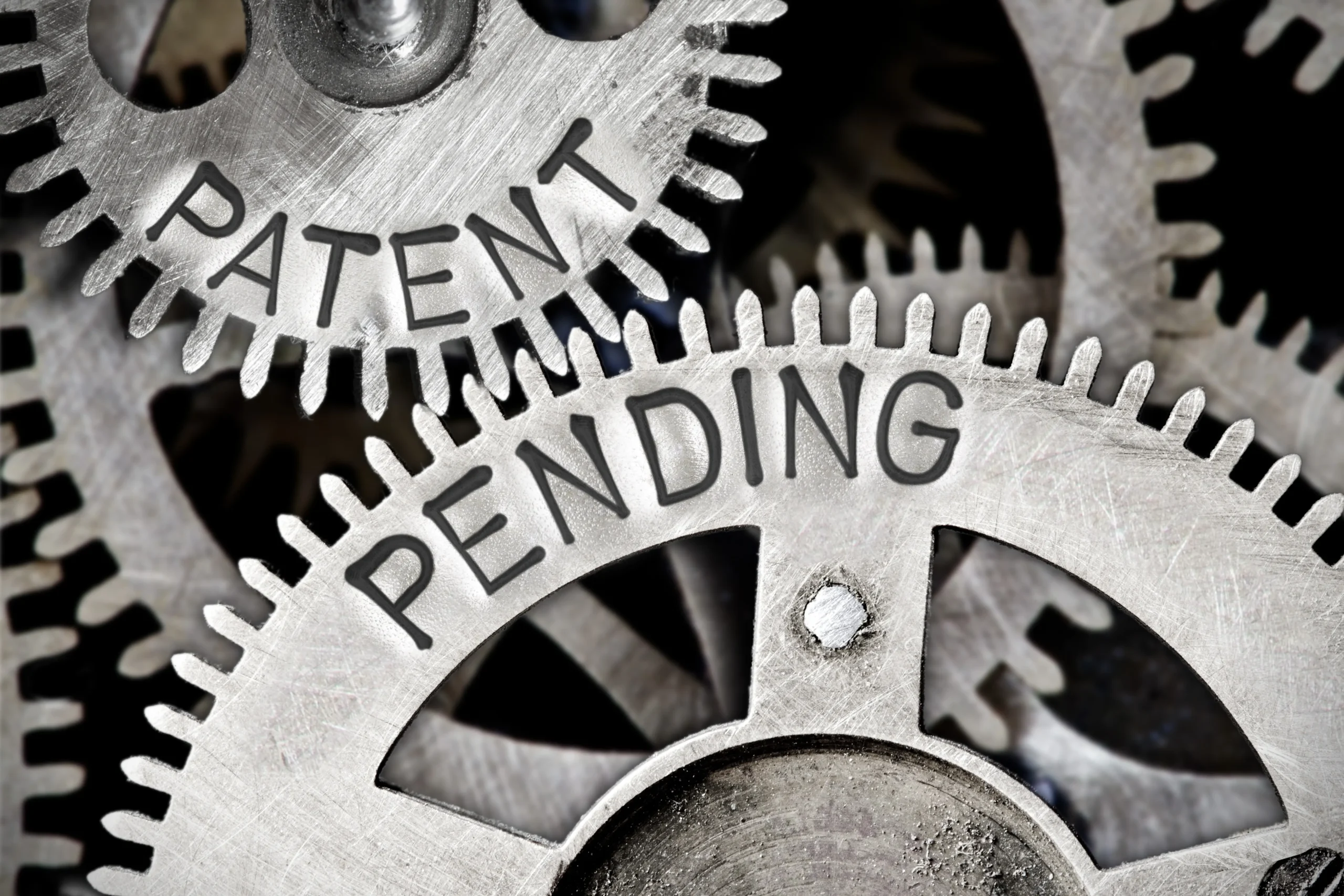 Close-up of interlocking metal gears, with the words "PATENT PENDING" clearly visible on two of them, symbolizing the complex machinery of intellectual property law and the concept of patent protection and potential government "march-in" rights.